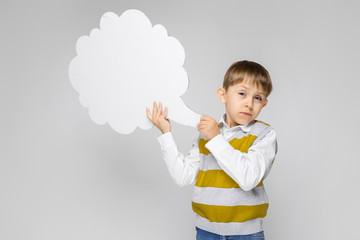 A charming boy in a white shirt, striped tank top and light jeans stands on a gray background. The boy is holding a white poster in the form of a cloud
