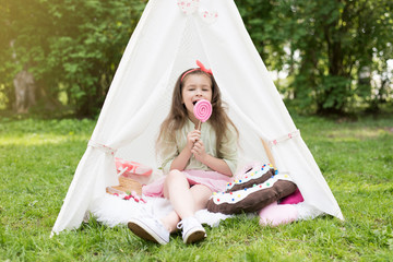 Little girl lying and playing in a tent, children's house wigwam in park. Happy mothers day. Child with big donuts and straw hat and lollipop. Summer, happy childhood concept. Cute emotions girl with © MartaKlos