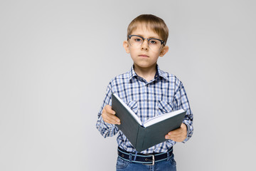 A charming boy with an inlaid shirt and light jeans stands on a gray background. The boy is holding a book in his hands. Boy with glasses