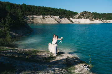 The bride in a beautiful dress with the groom in a light suit against the blue sky and blue water. Wedding couple standing on a sandy hill in the open air. A romantic love story.