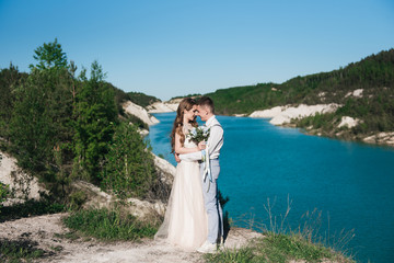 The bride in a beautiful dress hugging the groom in a light suit near the lake. Wedding couple standing on a sandy hill in the open air. A romantic love story. Azure-blue water on the horizon.