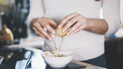 A young woman dressed in a white shirt is breaking an egg into a white bowl. A young woman is cooking in a modern kitchen.