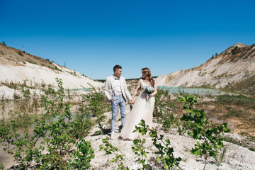 The bride in a beautiful dress holding hands with the groom in a light suit against the blue sky and blue water. Wedding couple standing on a sandy hill in the open air. A romantic love story.