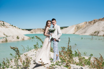 The bride in a beautiful dress hugging the groom in a light suit near the lake. Wedding couple standing on a sandy hill in the open air. A romantic love story. Azure-blue water on the horizon.