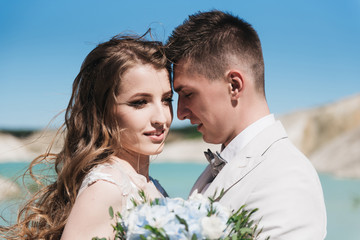 The bride in a beautiful dress hugging the groom in a light suit near the lake. Wedding couple standing on a sandy hill in the open air. A romantic love story. Azure-blue water on the horizon.