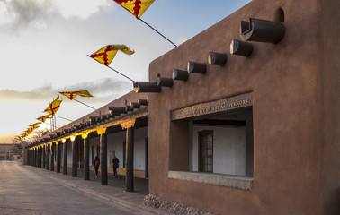 Obraz premium Flagi latające nad Pałacem Gubernatorów, Santa Fe Plaza, stolicą stanu Nowy Meksyk o zachodzie słońca w wiosenny wieczór. Struktura Adobe i historyczna hiszpańska siedziba rządu na południowym zachodzie.