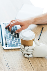 Fototapeta na wymiar robotic arm holding disposable coffee cup and human hand using laptop at wooden table