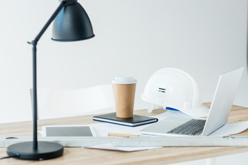 lamp, digital tablet, laptop, hard hat and disposable coffee cup on table