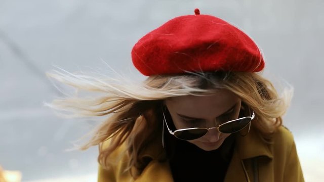 closeup outdoors portrait of attractive stylish woman wears red beret hat bright yellow jacket cool modern sunglasses looking at camera and pretty smiling sunny day fashion wind blowing in blonde hair