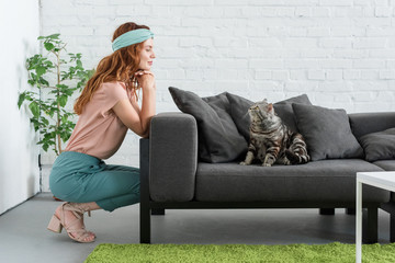 smiling young woman looking at her tabby cat while he sitting on couch at home