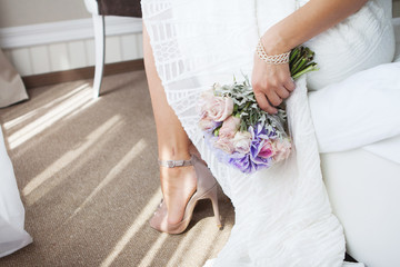 The bride sitting on a bed in wedding morning. Wedding dress and nude beige shoes. holding a wedding bouquet in her hands