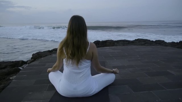 Zoom in with rear view of woman in white outfit sitting in lotus pose on stone dock near ocean and meditating in morning