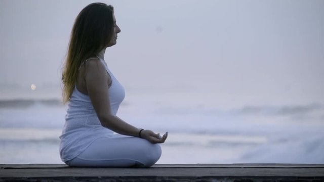 Side view of calm woman in white sitting in lotus pose and meditating near ocean