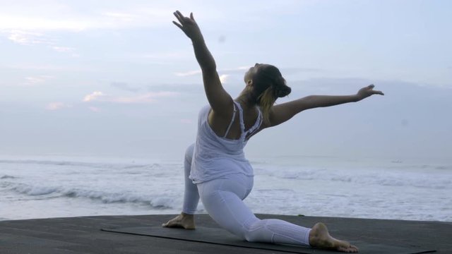 Medium shot of mindful woman in white doing low lunge yoga pose on coastline
