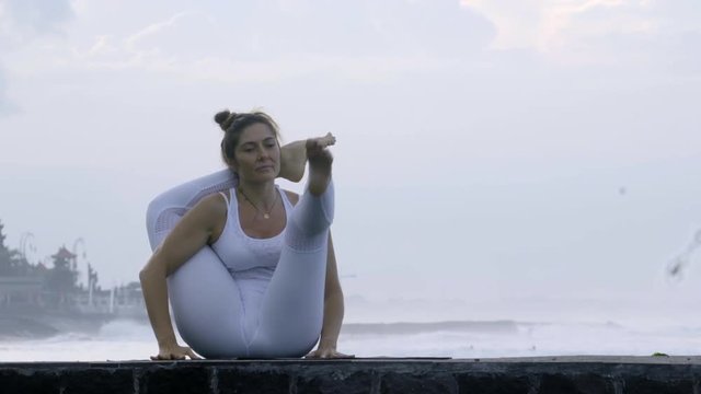 Strong flexible woman doing Eka Pada Sirsasana and forward fold pose on coastline in morning