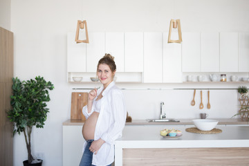 A pregnant woman is standing in the kitchen in jeans and a white shirt