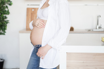 A pregnant woman is standing in the kitchen in jeans and a white shirt
