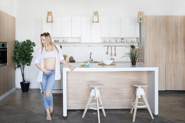 A pregnant woman is standing in the kitchen in jeans and a white shirt