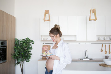 Pregnant woman in the kitchen holds a plate of cakes. A pregnant woman is standing in the kitchen in jeans and a white shirt