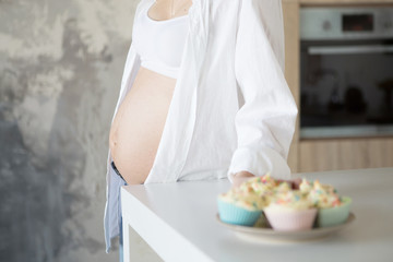 Obraz na płótnie Canvas A pregnant woman is standing in the kitchen in jeans and a white shirt. In the frame, a plate with cakes