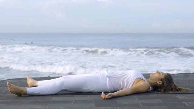 Side view of mature woman in white lying in Savasana pose and meditating on coastline near ocean