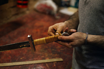 Smith makes leather winding of sword. Man is working in workshop. He fixes leather band on hilt of sword.