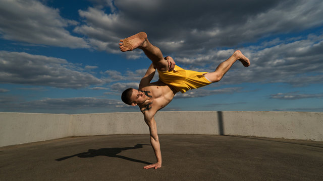 Tricking on street. Martial arts. Man performs blow with support of his hand barefoot. Shooted from bottom foreshortening against sky.