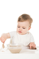 baby boy eating porridge by spoon and sitting in highchair isolated on white background