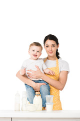 young mother with little son in hands at table with bottle of milk, plastic containers, bowl and baby bottles isolated on white background
