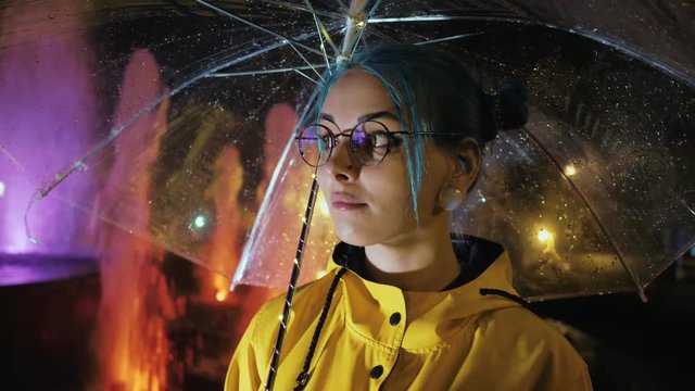 Young pretty girl with blue dyed hair in yellow raincoat and with transparent umbrella stands near fountain. Night illumination of city. Portrait of stylish hipster with glasses.