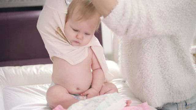 Mom undressing baby. Little baby in diaper strip clothes. Infant going for a walk. Young mother take off cloth from child. Baby unclothing on bed