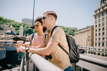 Fototapeta premium multicultural happy couple of tourists with coffee cups looking at view from bridge