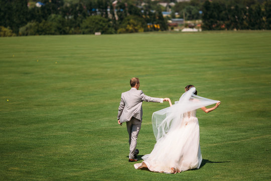 Rear view, newlyweds are walking along the green field of the golf club on a wedding day. The bride and groom in wedding dresses are holding hands. Man in a business suit in gray and beautiful woman