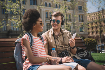 stylish couple with camera, headphones and smartphone sitting on bench
