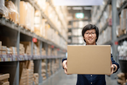 Young Asian happy man carrying cardboard box between row of shelves in warehouse, shopping warehousing or working pick and packing concepts