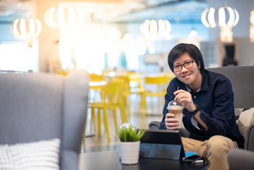 Young Asian happy businessman dressed in casual style drinking coffee working with digital tablet in cafe. Freelance lifestyle or digital nomad concepts