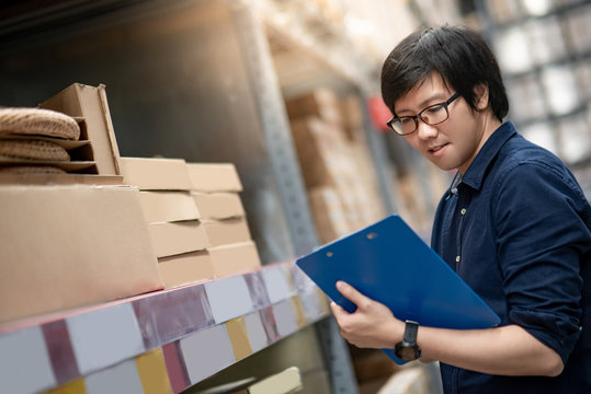 Young Asian man doing stocktaking in warehouseYoung Asian man doing stocktaking of product in cardboard box on shelves in warehouse by using clipboard and pen. physical inventory count concept