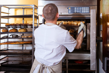 A male baker in white uniform and a beige apron bakes bread and takes cheese buns from an...