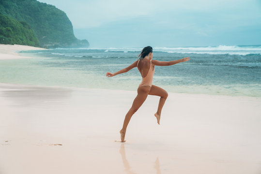A woman runs along the beach with white sand. Female athlete is engaged in sports in the morning near the ocean on the island. Model in a swimsuit is jumping.