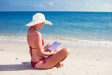 Fototapeta na wymiar Caucasian woman is reading a book on a sandy beach wearing a straw hat