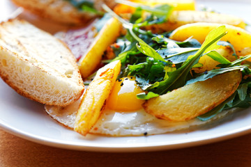 Rustic breakfast. Closeup fried eggs with fried slices of potatoes, bacon and fresh arugula on wooden table. Angle view. Shallow focus.