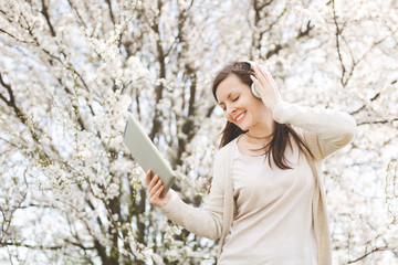 Young relaxed beautiful woman in light casual clothes with headphones listening music holding tablet pc computer in city garden or park on blooming tree background. Spring flowers. Lifestyle concept.