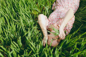 Young charming woman in light patterned dress lying on grass keeping hands near face looking up resting in sunny weather in field on bright green background. Spring nature. Lifestyle, leisure concept.