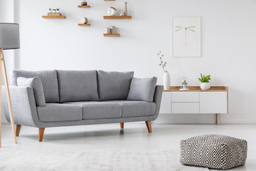 Patterned pouf and grey couch in minimal living room interior with poster above cupboard. Real photo