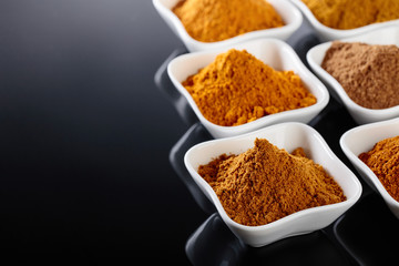 Small white ceramic bowls of Indian spices on black reflective background.