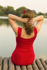 Fototapeta na wymiar Beautiful girl in red dress is sitting on wooden pier. Woman is holding her hair with red flower near the lake. Rustic and natural photo outdoors. Summer river and splashes of water in the evening.