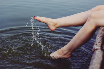 Beautiful girl in red dress is sitting on the wooden pier. Woman is dangling her feet into the lake. Rustic and natural photo outdoors. Summer river and splashes of water in the evening.