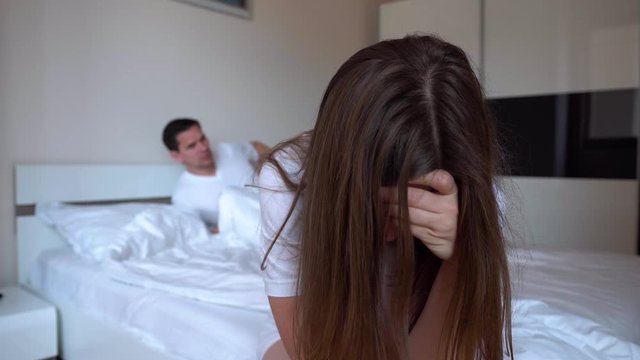 Woman Sits On The Edge Of The Bed After A Quarrel And Thinks About Something Sad Against The Background Of Blurred Indifferent Husband Going To Bed