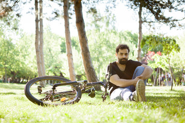 Fototapeta premium Young bearded man sitting on the grass in the park next to his bicycle