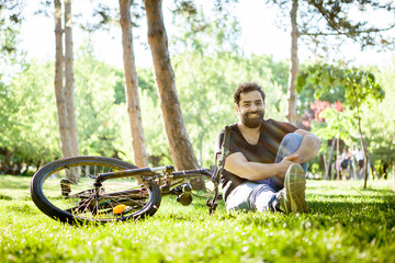 Young bearded man smiles to the camera sitting on the ground in the grass in the park. His bicycle is next to him on the ground